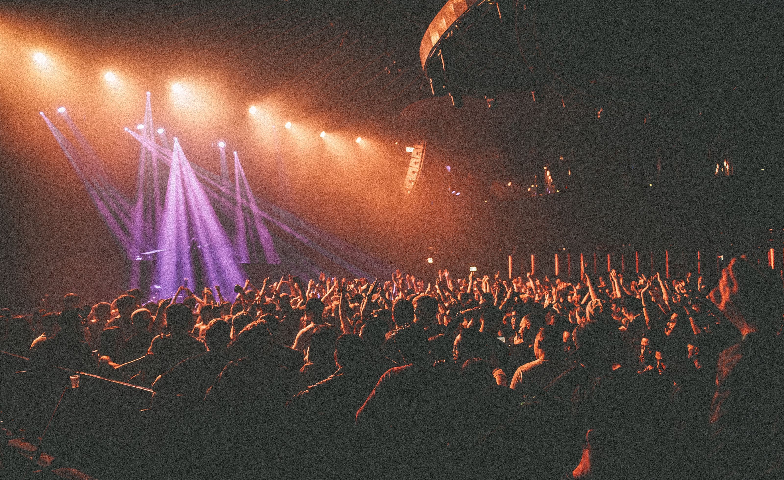 A packed concert crowd with hands raised under stage lights